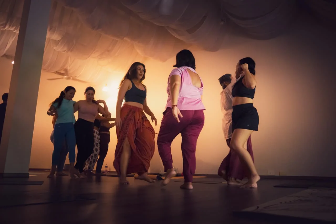 Grupo de personas en una práctica de danza en un salón con telas y luz cálida.